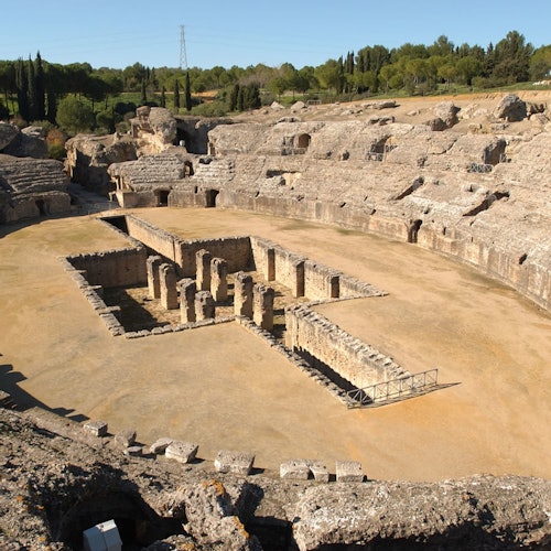 고대 원형 극장의 항공 뷰. 중앙에는 석재 폐허가 있고, 나무로 덮인 언덕에 둘러싸여 있습니다.