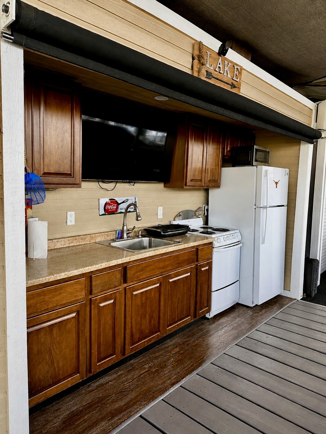 Outdoor kitchen area with wooden cabinets, a white refrigerator, and a stove under a "Lake" sign, featuring a mounted television and countertop sink.