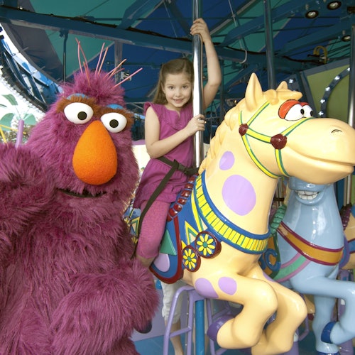 A child in a pink dress rides a carousel horse next to a large, furry, pink puppet character with an orange nose.