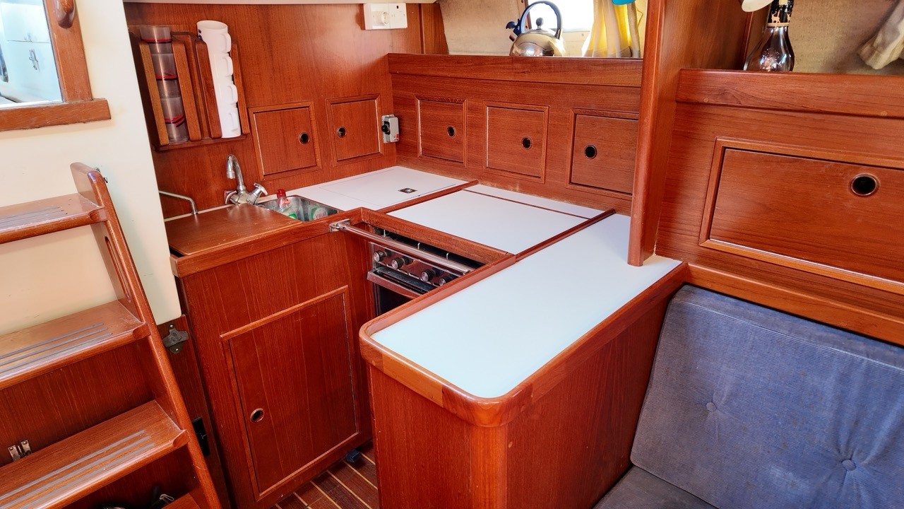 Contest 38 Ketch galley interior — teak cabinetry with multiple stowage lockers, gimballed oven and hob with fiddle rails, stainless steel sink, white laminate worktops, companionway steps, and steel kettle on shelf — offshore bluewater cruising yacht galley