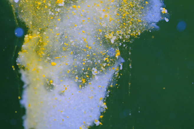 Yellow and white powder dispersing in green liquid, creating a cloud-like swirl with scattered bright particles.