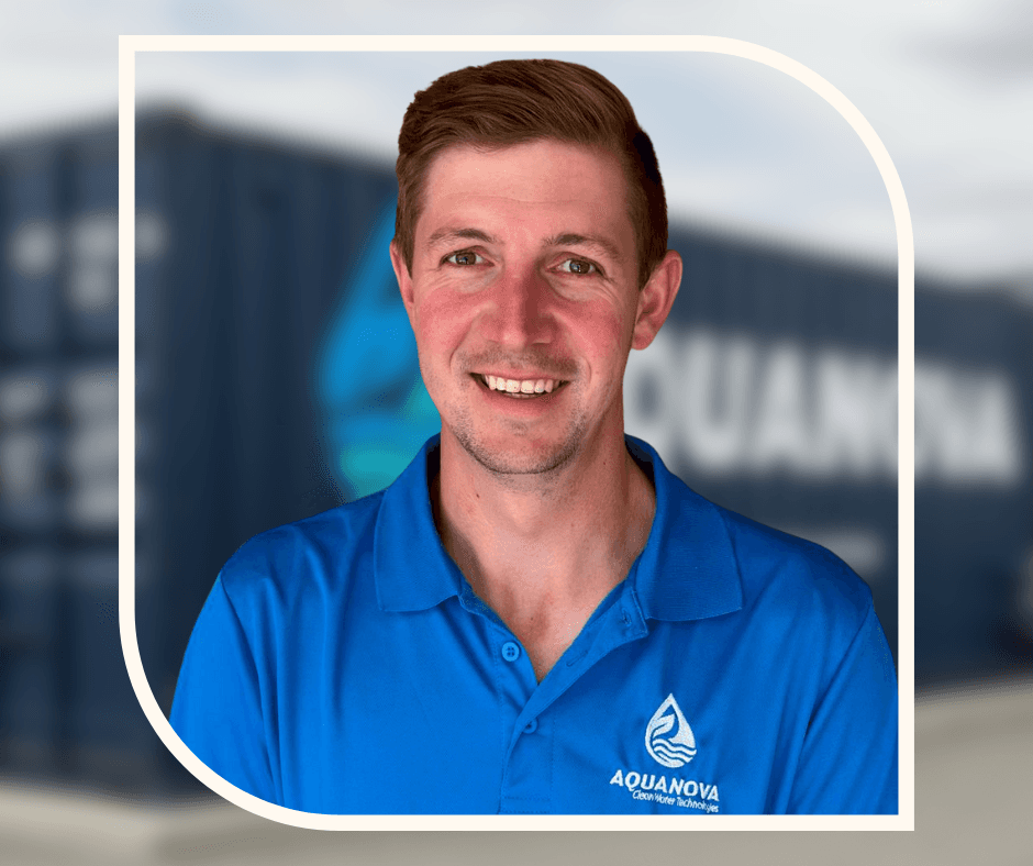 A smiling man with curly hair is sitting at a desk, looking at the camera, with a laptop and a lamp in the background.