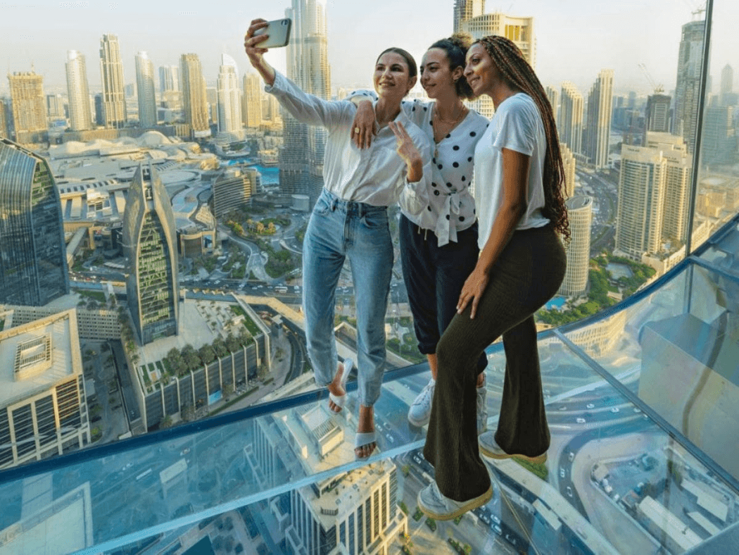 3 young women taking a selfie as they stand on the glass walk of the Sky View where you can see Dubai behing and below them.