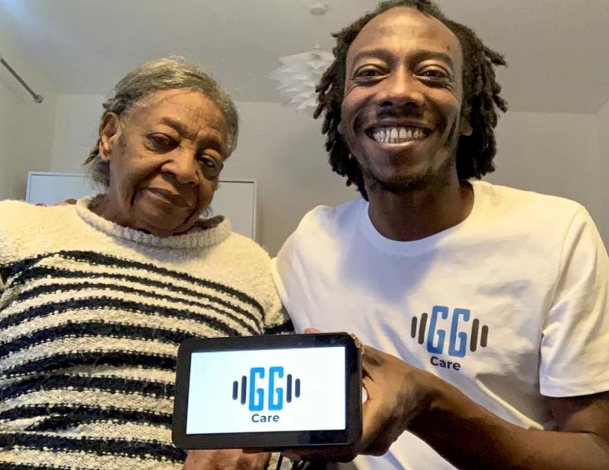 A photo of an elderly woman wearing a striped jumper stands next to a man with short black dreadlocks wearing a white t-shirt with the GG Care logo on it. The man is holding an amazon alexa device with the GG Care logo on the screen