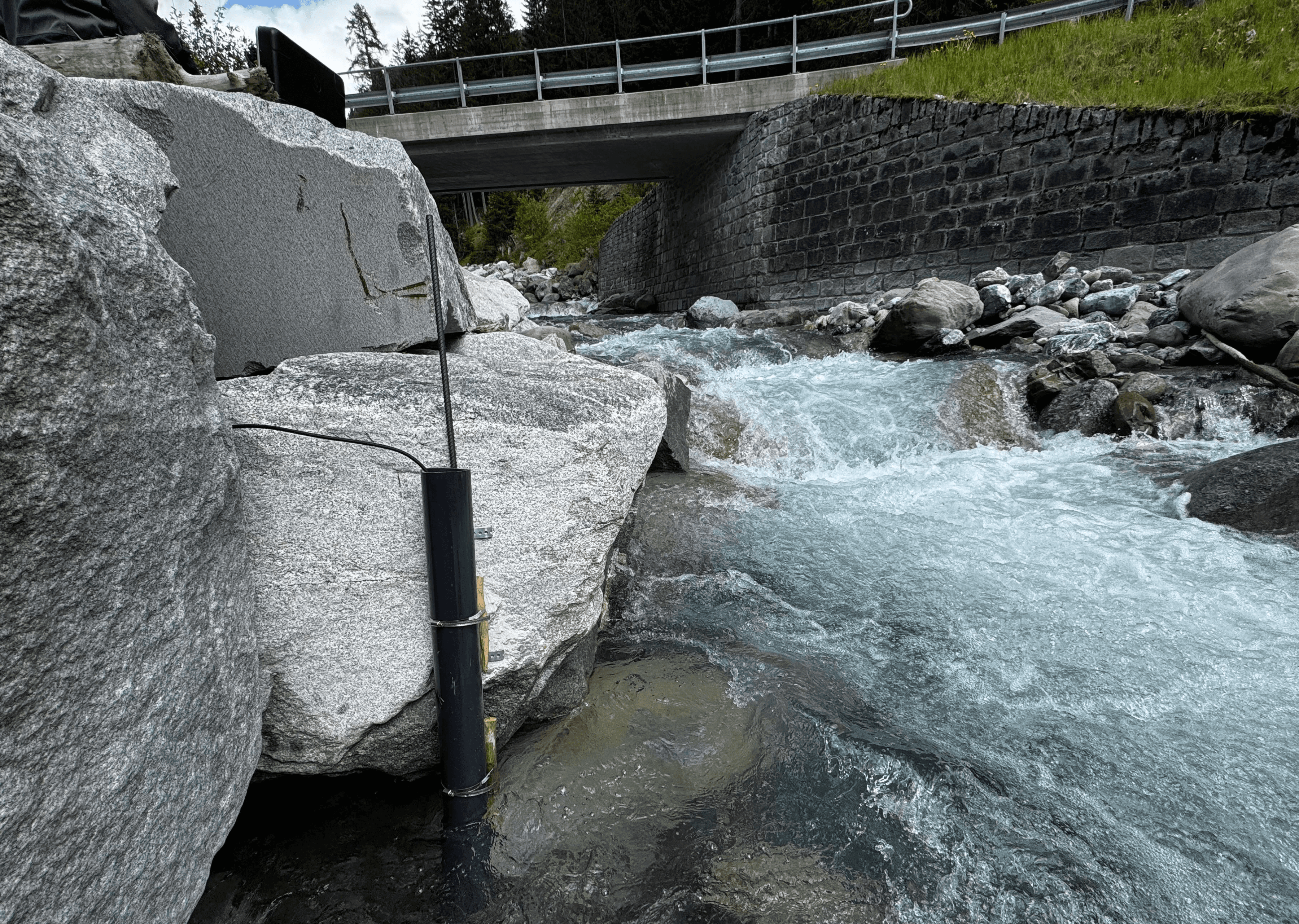 River monitoring device installed among rocks in a flowing stream beneath a bridge.