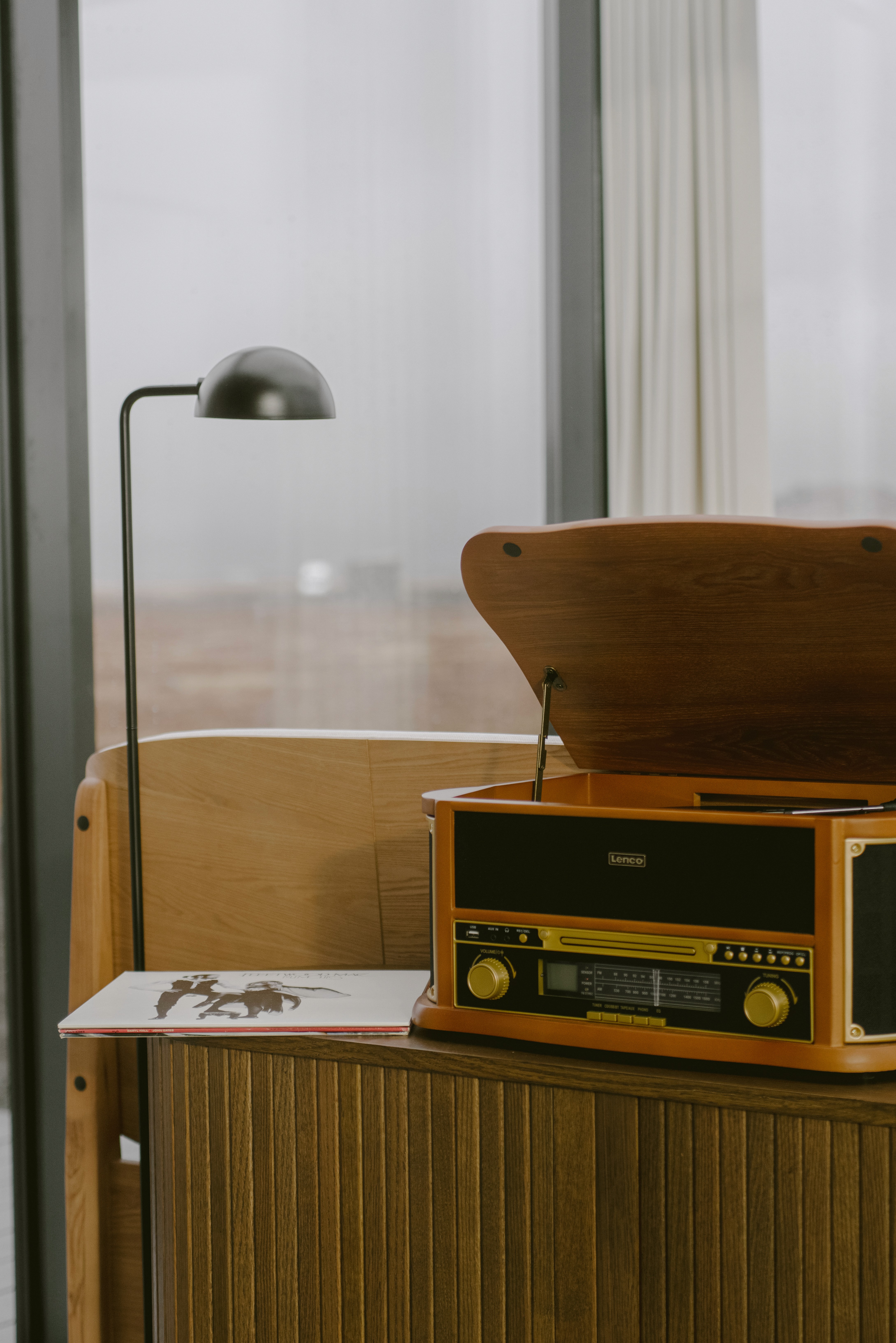 Vintage-style record player and radio on a wooden cabinet beside a reading lamp, set against a glass wall with views of an open landscape.