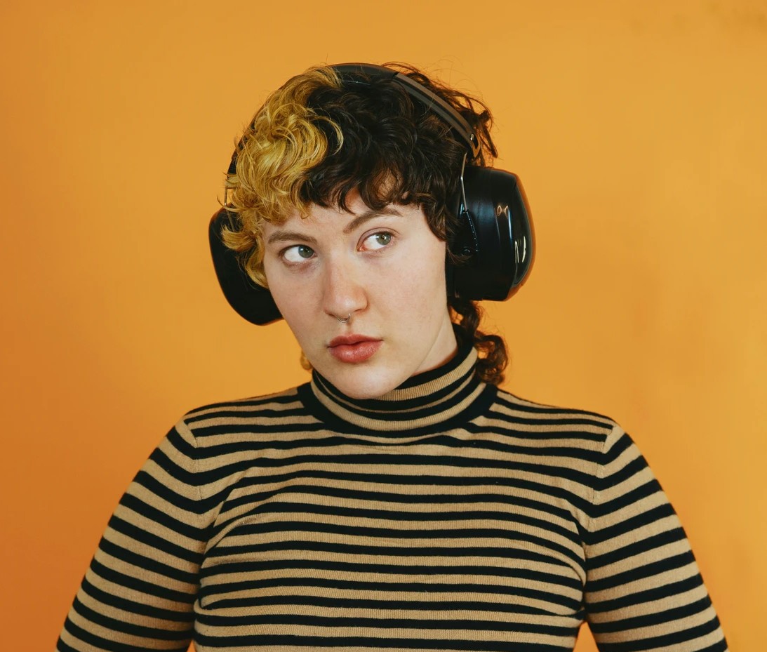 A young person with curly hair wearing large headphones and a striped top, against an orange background.