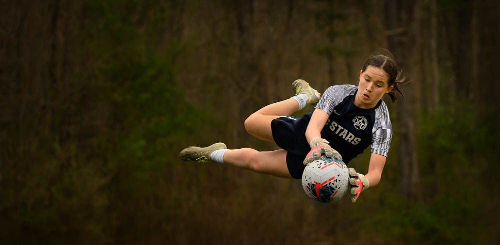 A female youth goalkeeper leaps through the air holding the ball against a dark background, showcasing agility and athleticism.