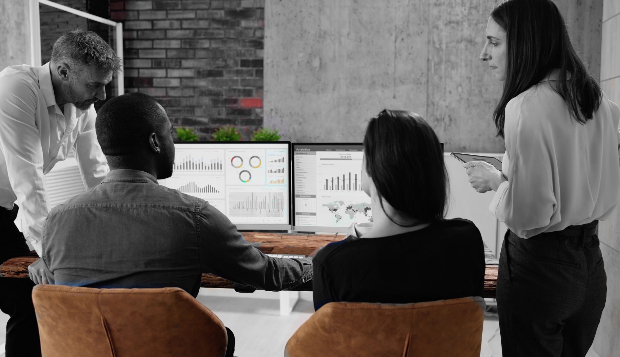 Black and white photo of a smaal room. At the room centre is a table. the are 2 clients sitting at the table with 2 computer screens showing graphs in front of them. They are involved in a discussion with 2 consultants, male and female, positioned on each of the table' sides.