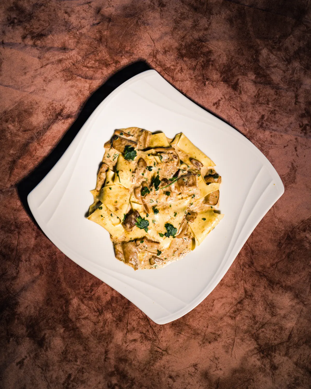 Three people in casual outdoor attire stack firewood into a rustic stone oven amidst an olive grove, with a sunlit house in the background, capturing an authentic rural lifestyle scene.