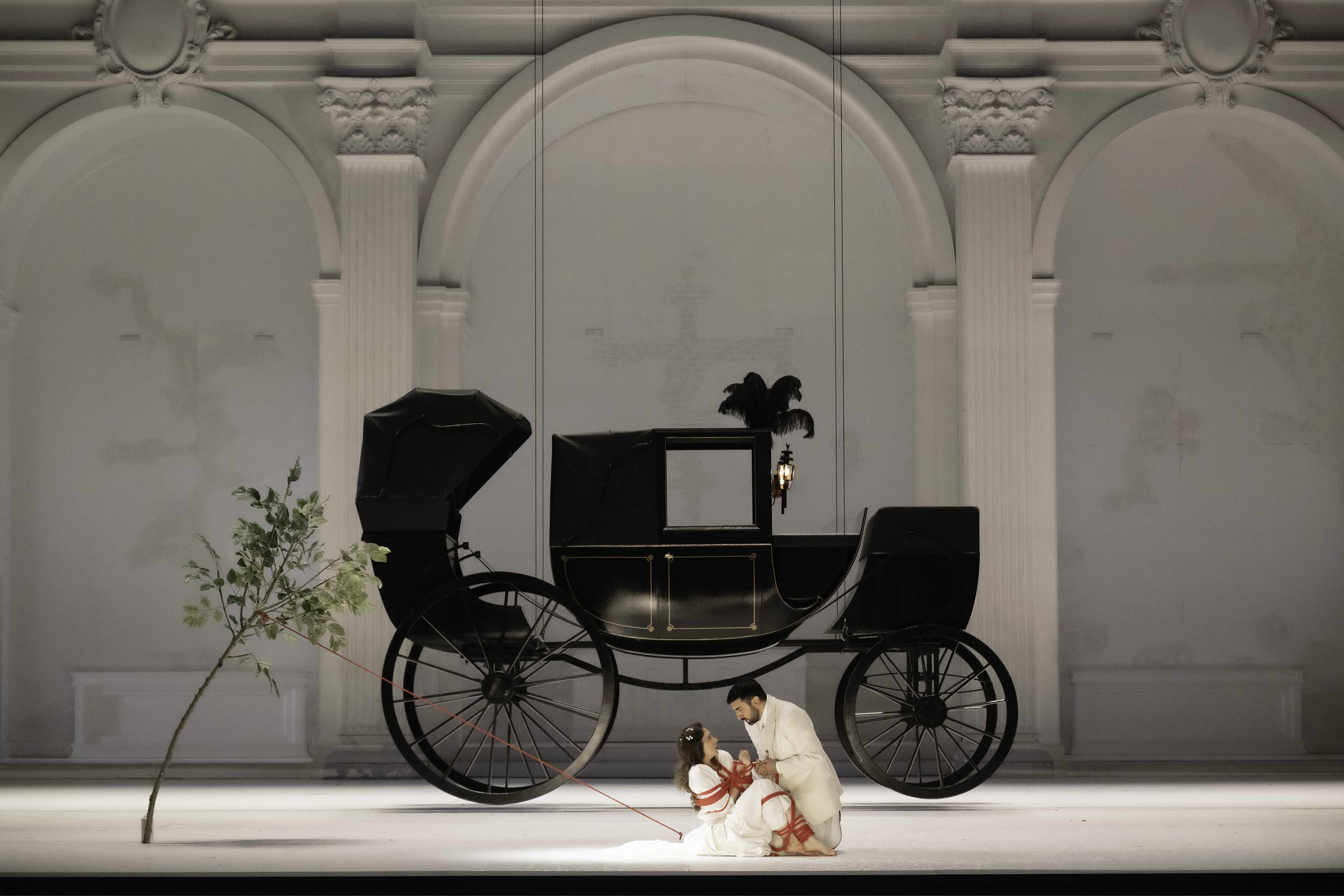 A girl in a white dress is being tied with red rope by a man in a white suit in front of an elegant black old-fashioned carriage.
