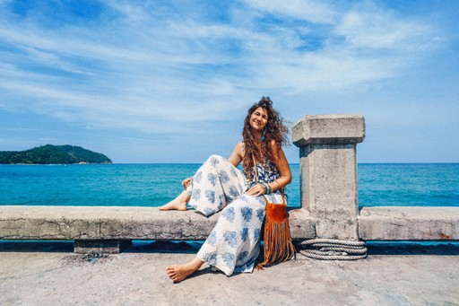 A person sitting on a ledge by the sea, dressed in white attire, with a scenic ocean and sky backdrop.