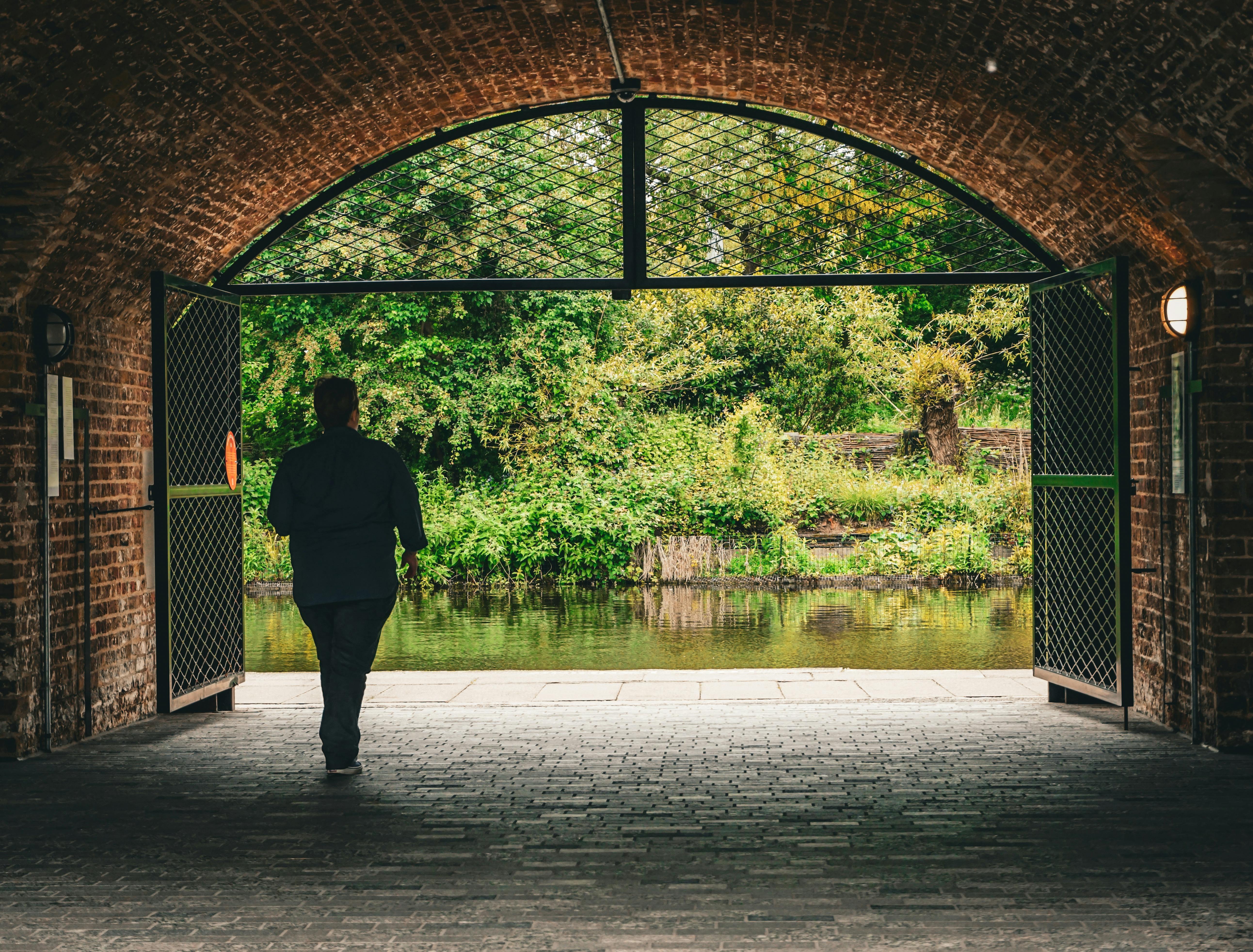 A wide opening in a brick building with open gates Outside is the lush forest type area and a lake a single person is silhouetted in the open doorway walking towards the forest