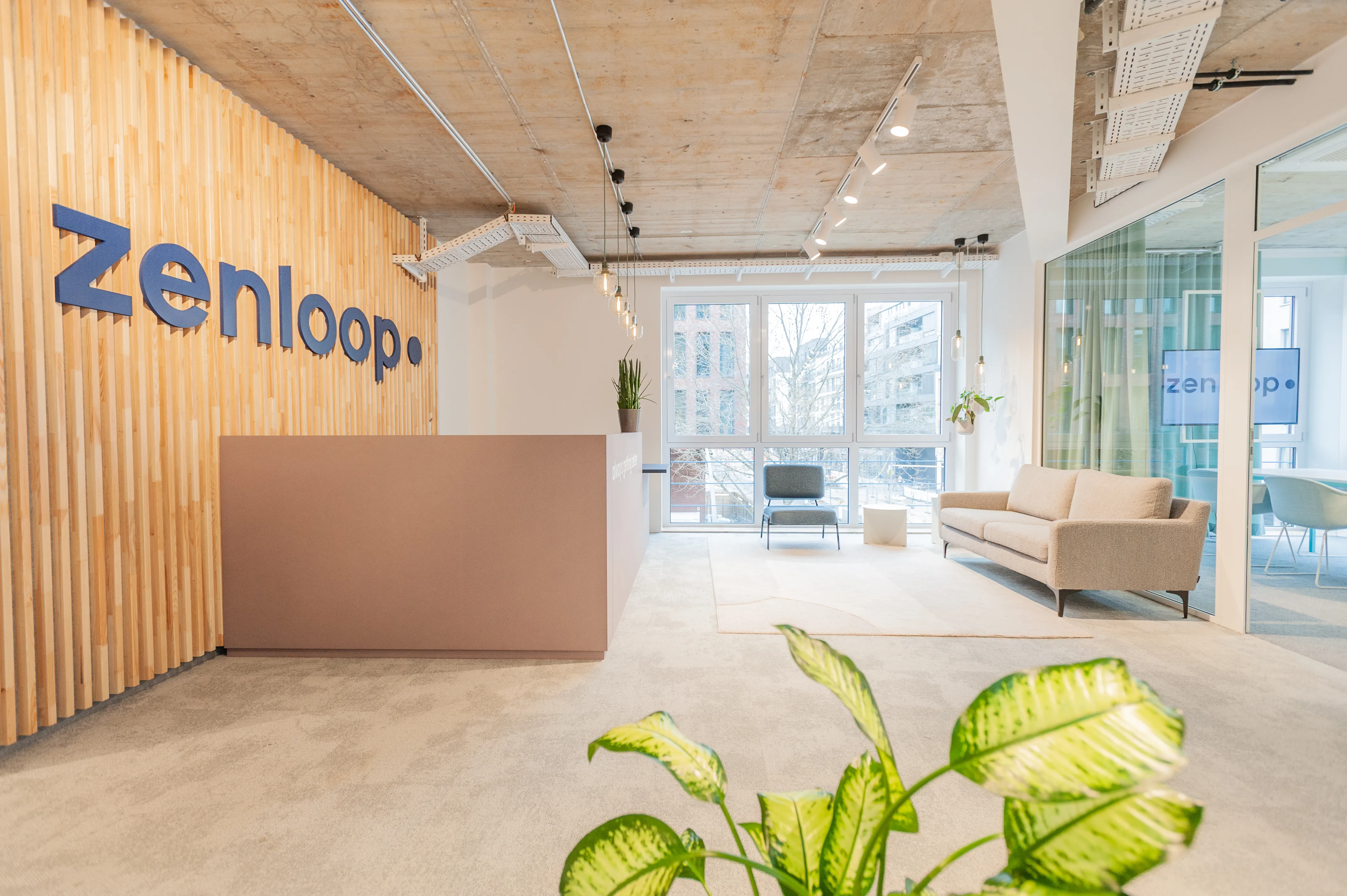 Waiting corner at ZENLOOP with blue corduroy chairs around white bistro tables, an oak sideboard with drawers, a cornflower-blue wall niche with plants and the navy logo on white.