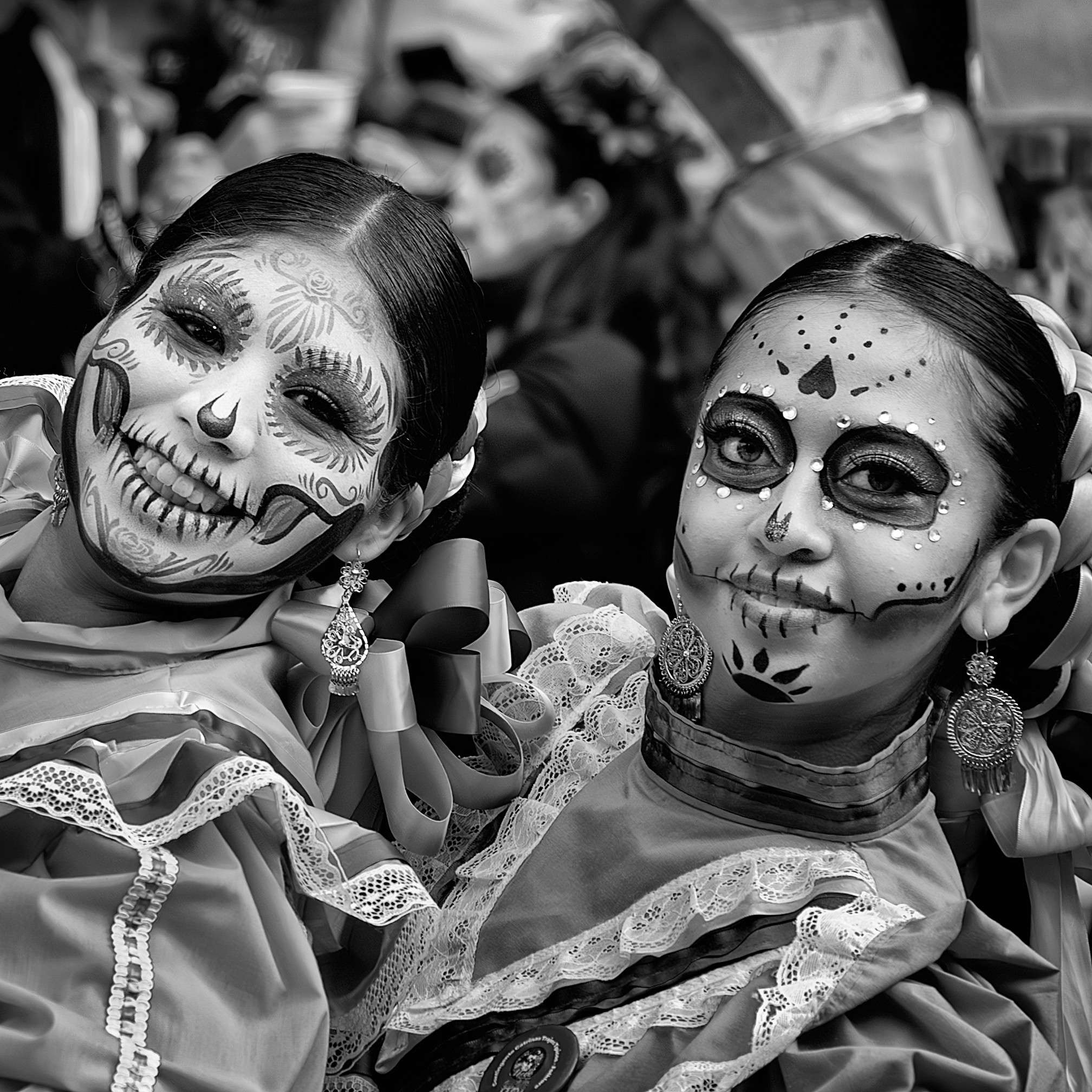 Black and white portrait of two women in traditional folkloric dresses with elaborate Day of the Dead face paint, smiling and posing during a festive celebration.