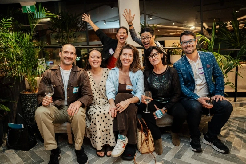 A group of seven people sitting and standing cheerfully at a social gathering, surrounded by vibrant plants, with some holding wine glasses, on a patterned tiled floor inside a modern, warmly-lit venue.