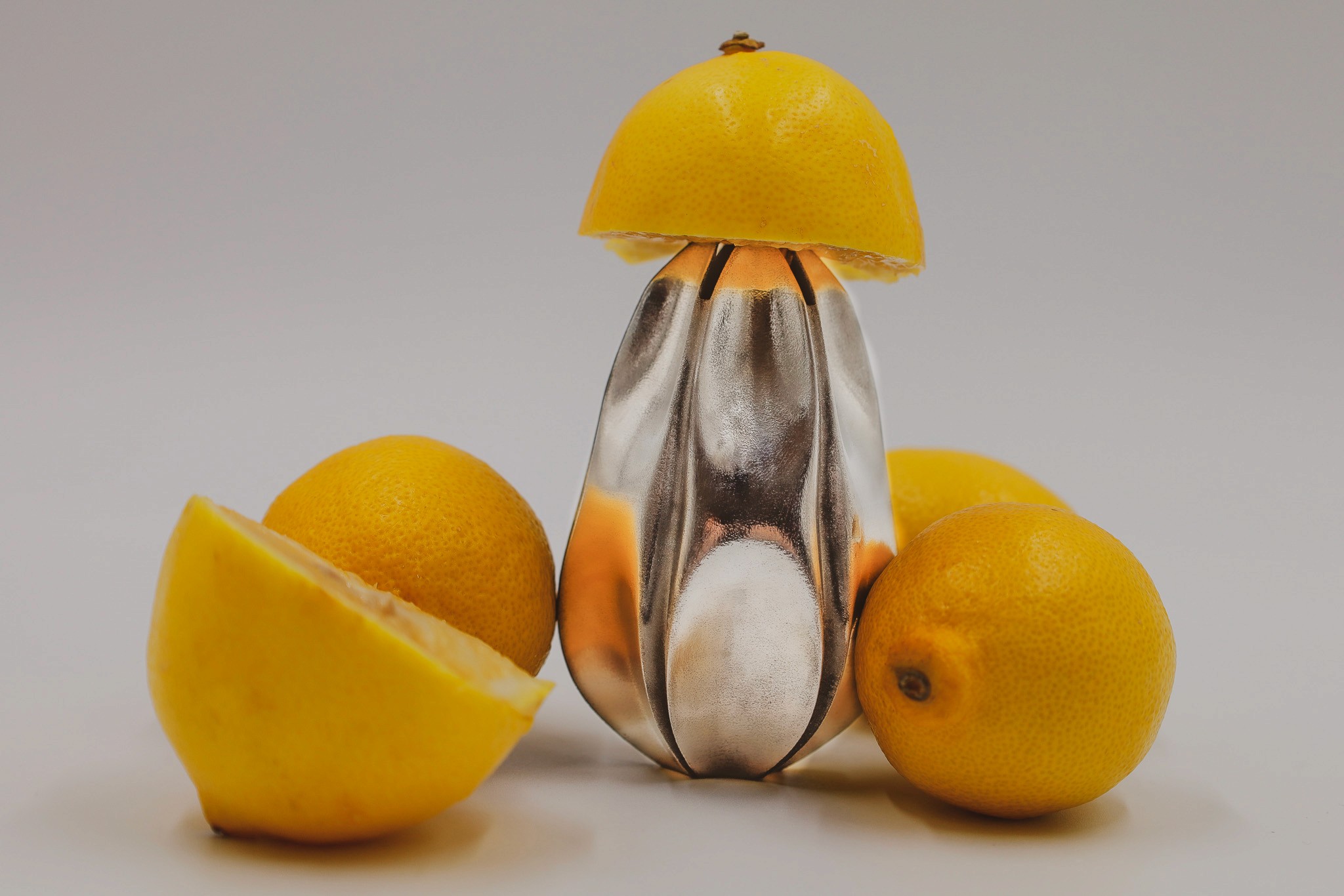 A glossy, reflective squeezer surrounded by whole and sliced lemons, with one half of a lemon placed on top as if it were a hat.