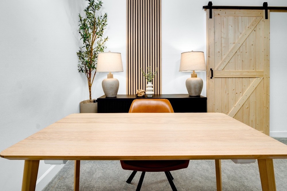 Book The Office “Minimalist office setup with a wooden desk and two chairs, accented by a sliding barn door, console table with lamps, and indoor plants for a warm, professional atmosphere.”