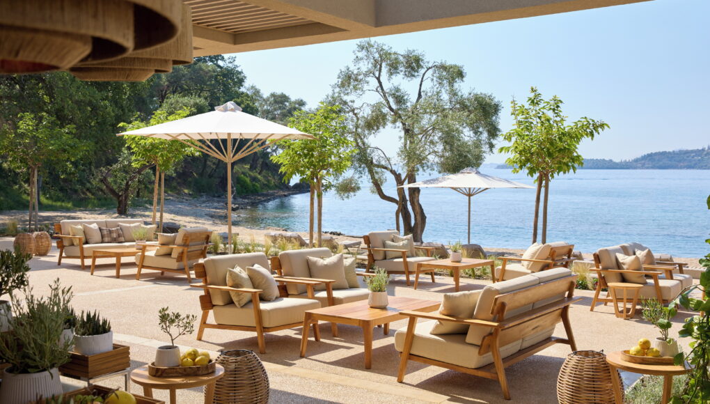 Gemütlicher Außenbereich einer Hotel-Lounge mit beigen Polstermöbeln, Holztischen und Sonnenschirmen direkt am Meer unter schattenspendenden Bäumen.