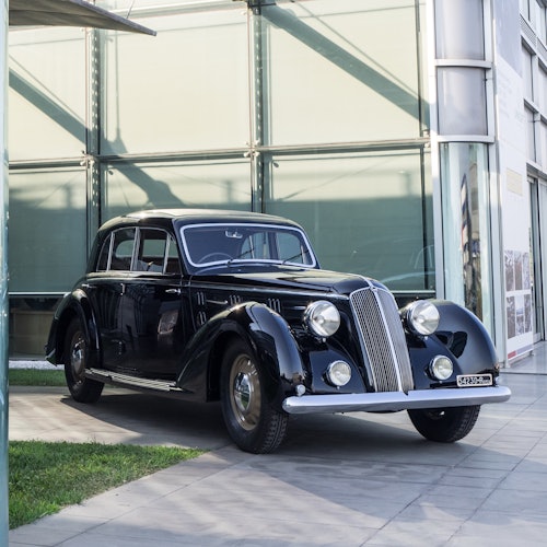 یک خودروی کلاسیک مشکی با چراغ‌های جلو گرد بر روی سطح کاشی‌کاری شده نزدیک یک ساختمان شیشه‌ای مدرن پارک شده است.