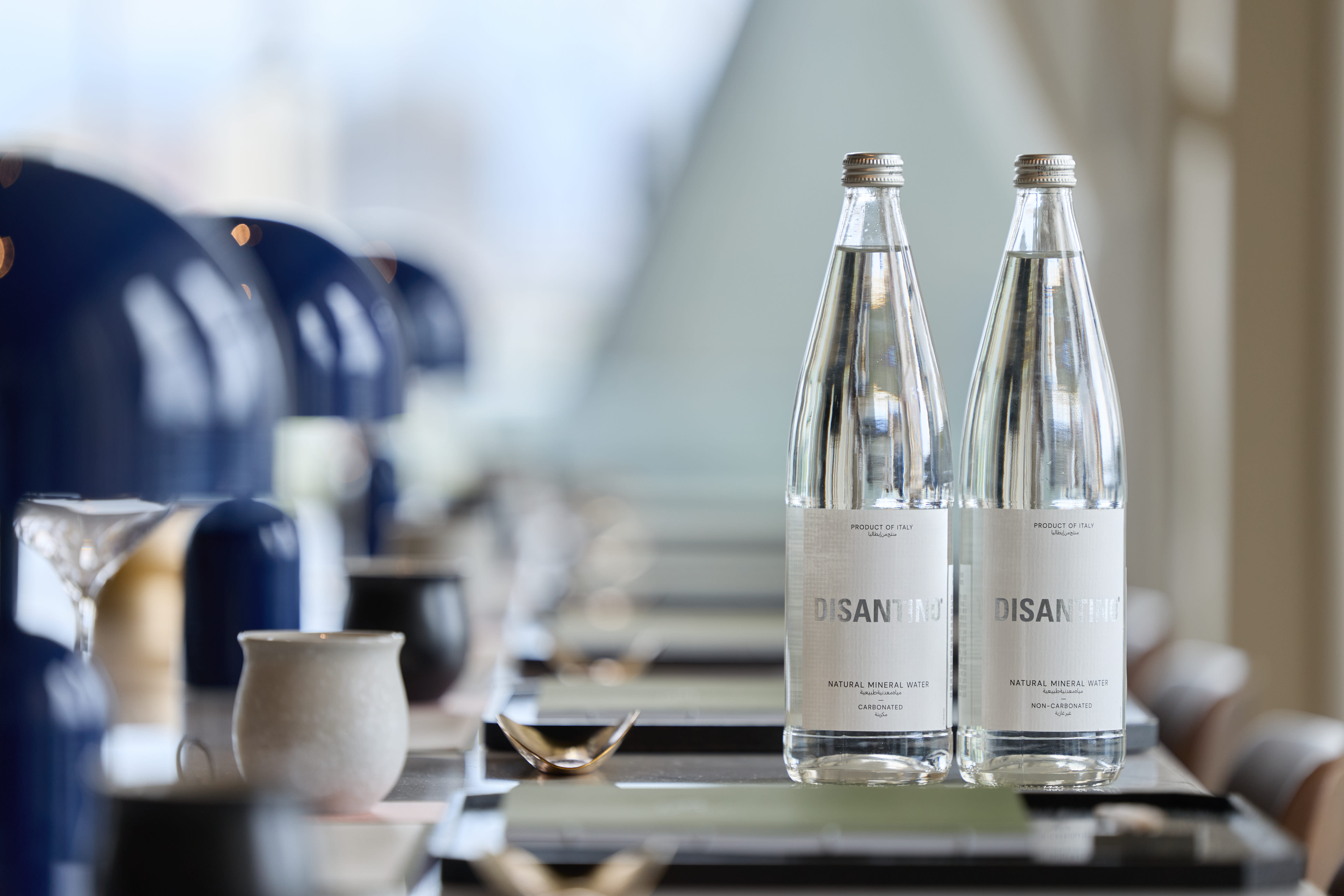 Two elegant glass bottles of DISANTINO Natural Mineral Water—one carbonated and one non-carbonated—standing on a sleek, reflective dining surface in a sophisticated, blurred interior setting.
