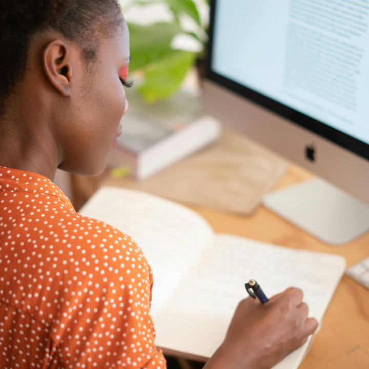 A person in an orange polka dot shirt writes in a notebook on a desk with a computer displaying text. Green plant and book in the background.