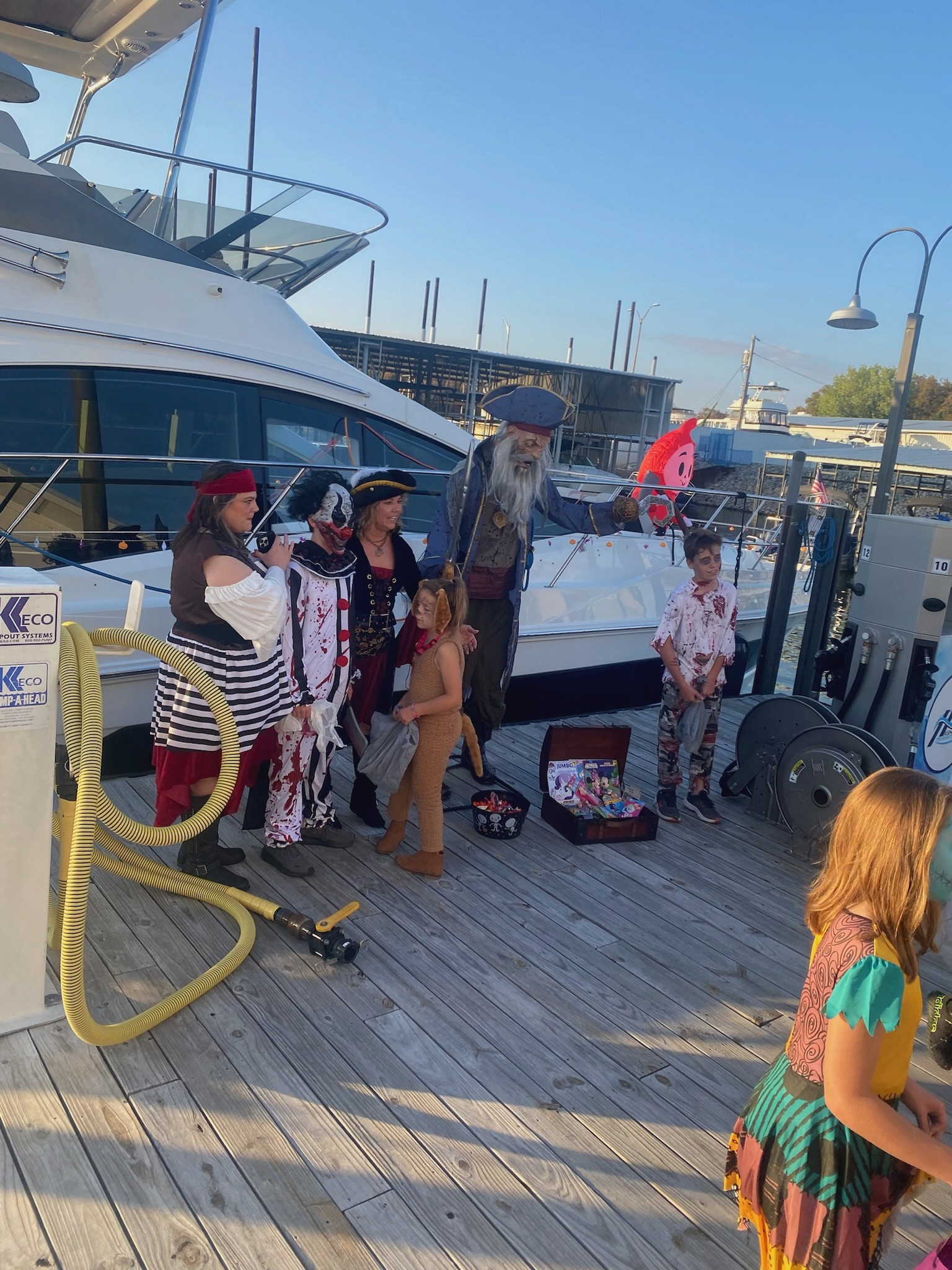 A group of people, dressed in pirate-themed costumes, stand on a wooden dock beside a large boat during a sunny day, with a treasure chest filled with candy nearby, enhancing the festive Halloween atmosphere at the marina.