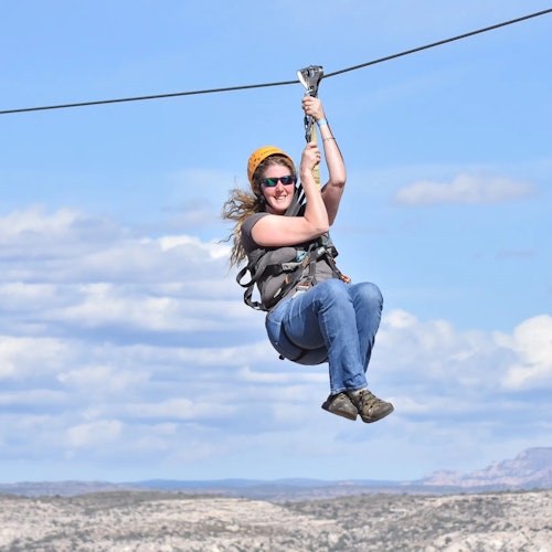 Усмихнат човек, носещ каска и слънчеви очила, се спуска по въже над скалист пейзаж под синьо небе с разпръснати облаци.