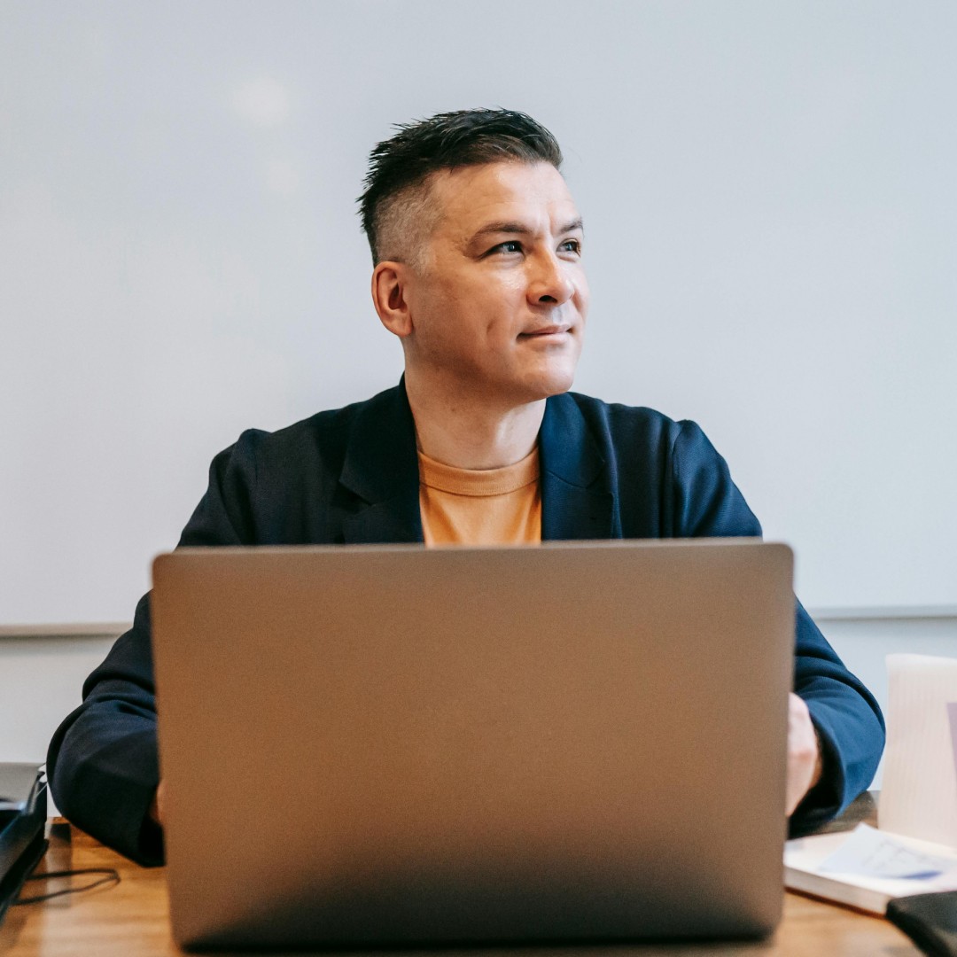 Two women are standing side by side against a beige wall, smiling confidently, one wearing a green shirt and the other in a dark blazer, with soft lighting casting gentle shadows.