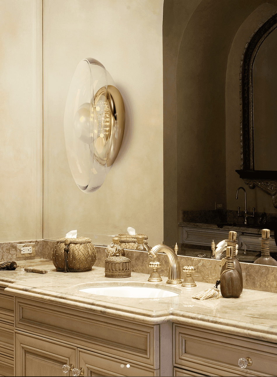 Luxury bathroom interior featuring a sculptural glass and bronze wall sconce above a marble vanity with brass fixtures and warm ambient lighting