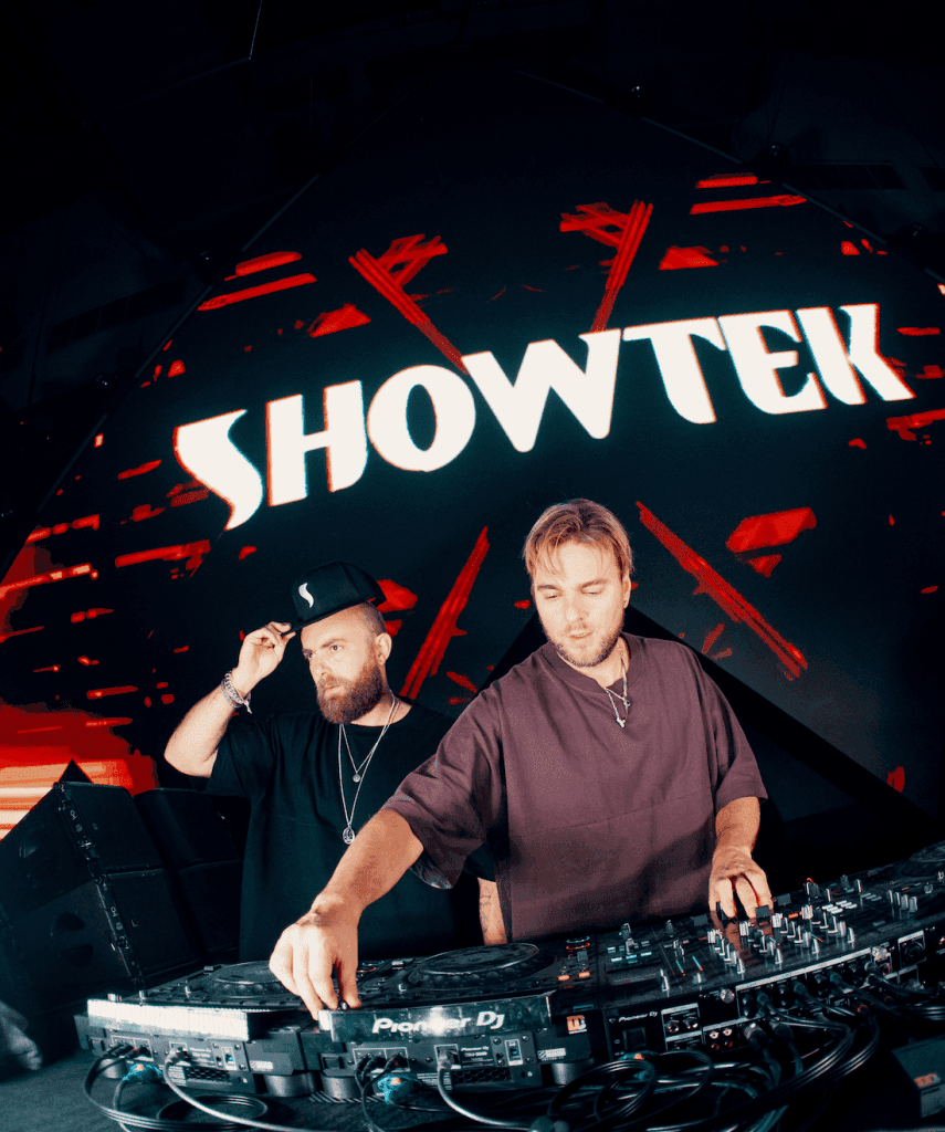 Two DJs performing on stage with a large screen displaying the name 'SHOWTEK' in the background. The atmosphere is vibrant with colorful lighting.