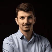 Daniel Antoni, Founder & CEO der VisioSEO GmbH, Portrait eines Mannes mit verschränkten Armen im hellblauen Hemd vor moderner Büroarchitektur mit Glasfassade.