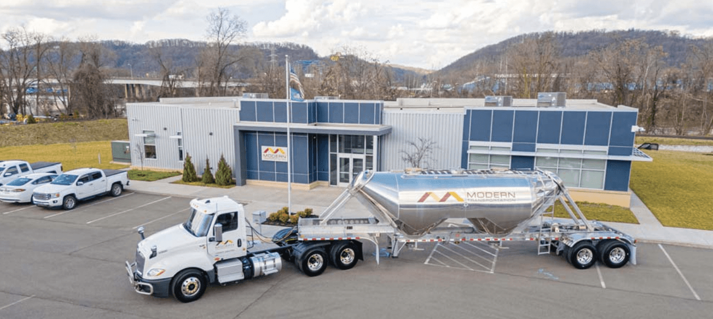 A white and blue heavy chemical tanker truck is parked in the yard in front of the company building, also in the same neighborhood. In front of the company, you can see the American flag and the “Transporte Moderno” company logo.