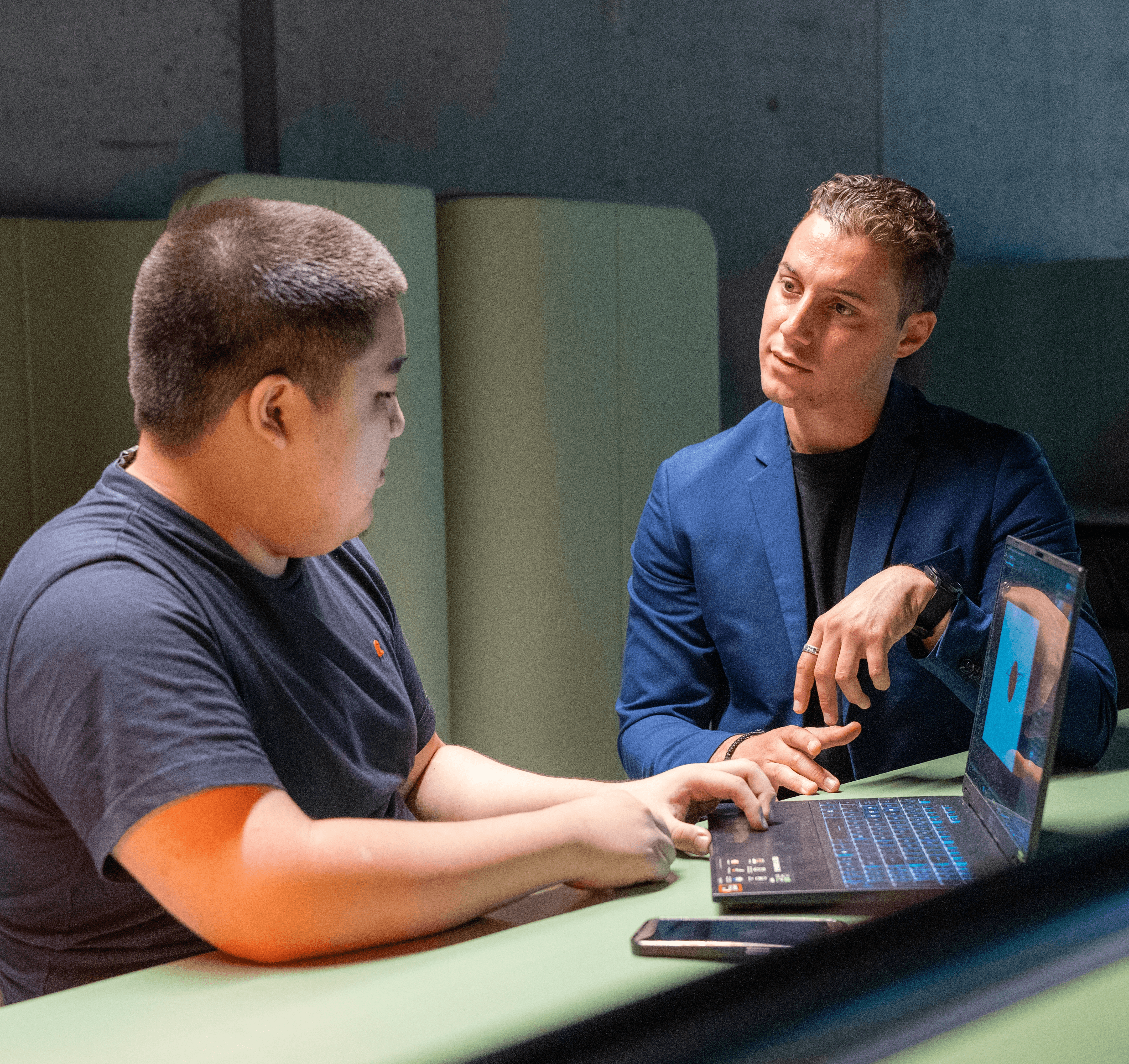 A young person with curly hair smiles at the camera while sitting in front of a laptop against a dark background.