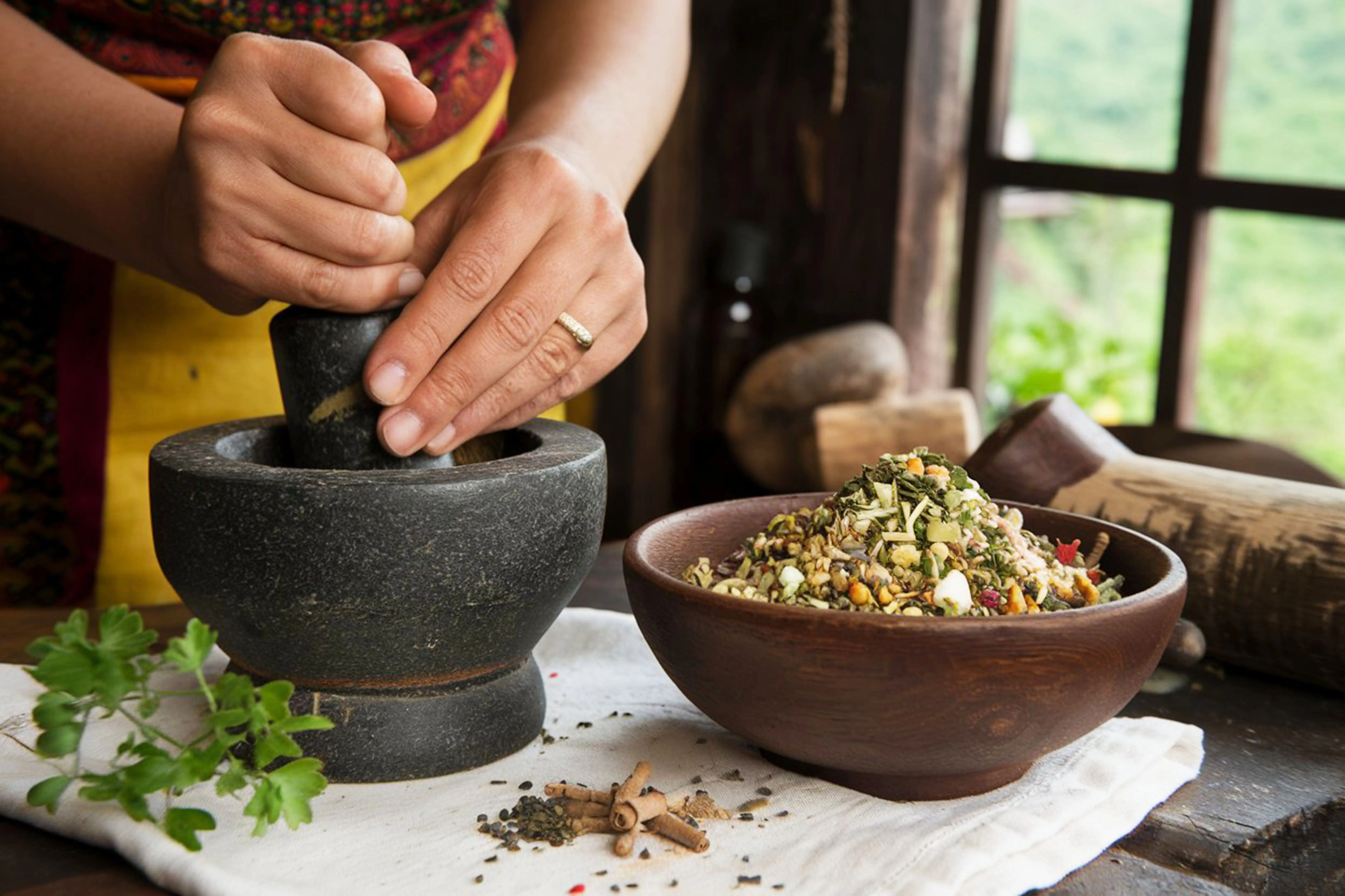 Close-up of Ayurvedic practitioner preparing herbal medicines using a stone mortar and pestle, with a bowl of traditional healing ingredients, representing authentic wellness treatments at Au Revoir Resort.
