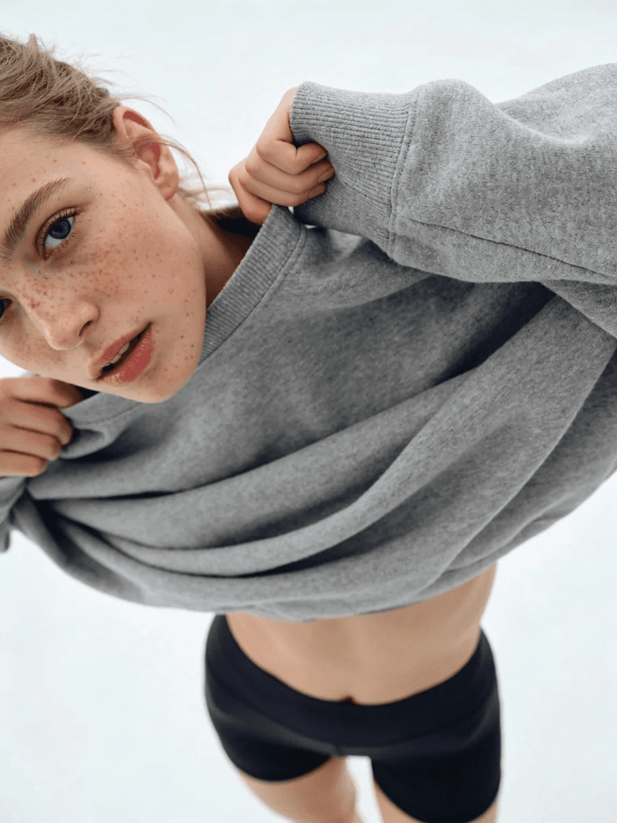 Close-up of a person in a gray sweatshirt and black shorts, lifting the sweatshirt slightly, with a white background.