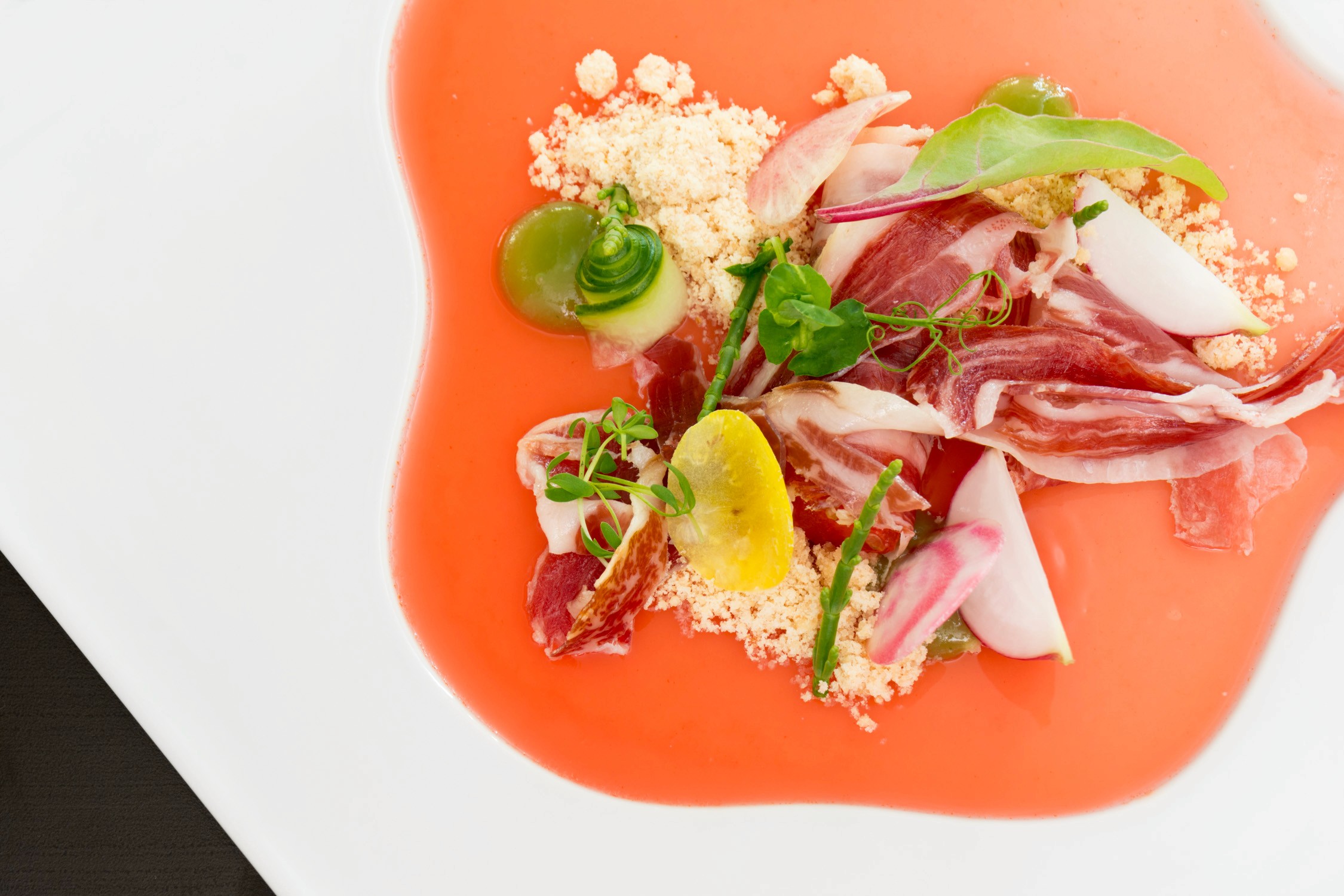 A gourmet appetizer featuring thinly sliced prosciutto and avocado on a vibrant orange sauce with herbs and grains.
