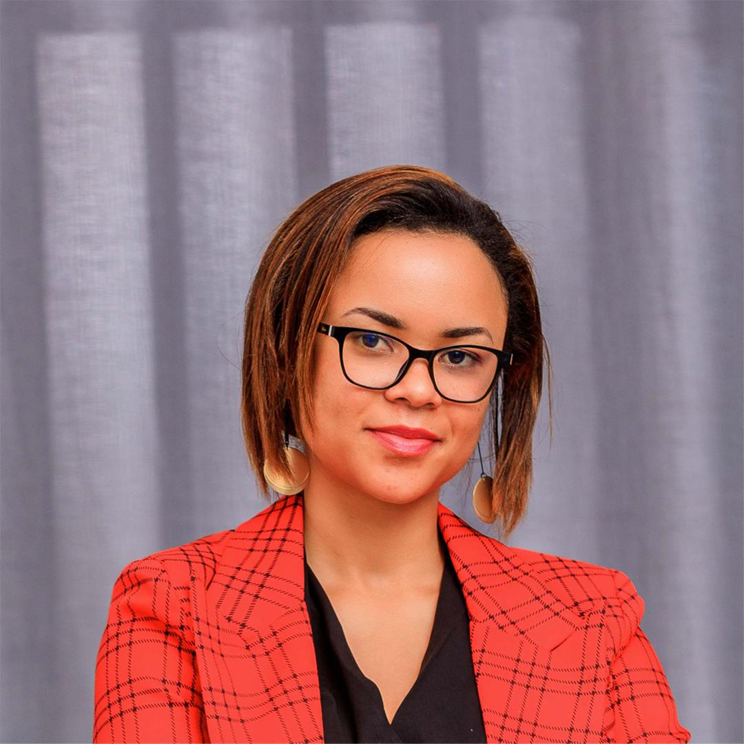 A young person with curly hair smiles at the camera while sitting in front of a laptop against a dark background.