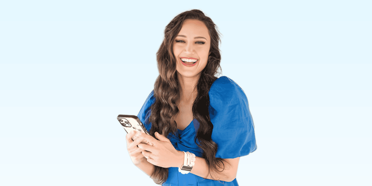 Sonia Begonia smiling while holding her phone, dressed in a blue outfit, radiating confidence and style in a joyful moment.