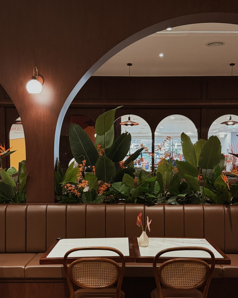 Interior view of a restaurant with an arched opening, featuring brown leather seating, two tables with white marble tops, and decorative plants. Soft lighting and a glimpse of the dining area through a window can be seen in the background.