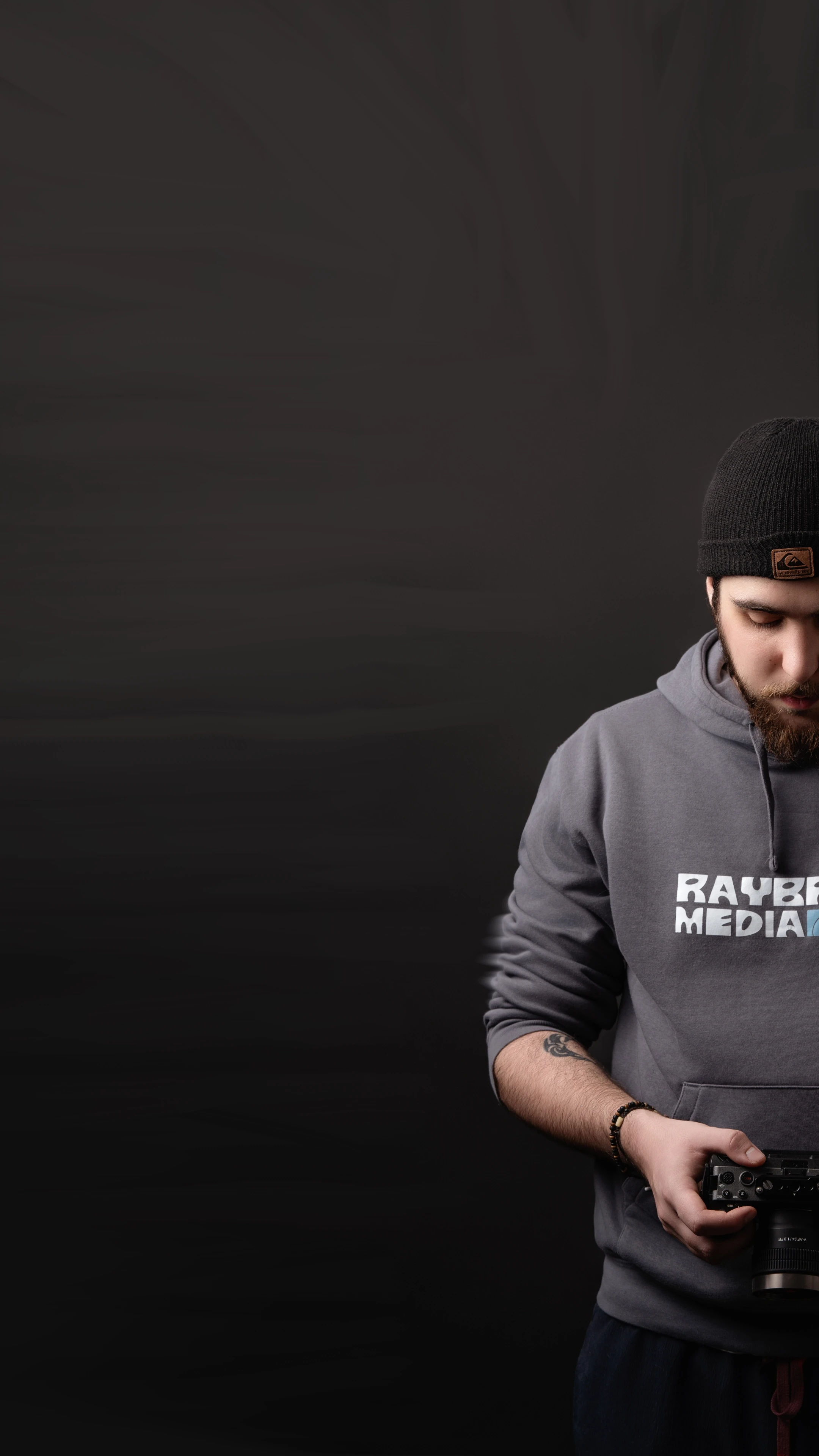Videographer wearing Raybrand Media hoodie adjusting a camera against a dark background, representing professional video production, content creation and personal brand identity