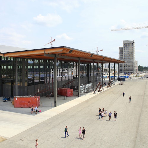 Pessoas caminham em um amplo calçamento ao lado de um prédio moderno coberto de vidro, com guindastes e arranha-céus ao fundo.