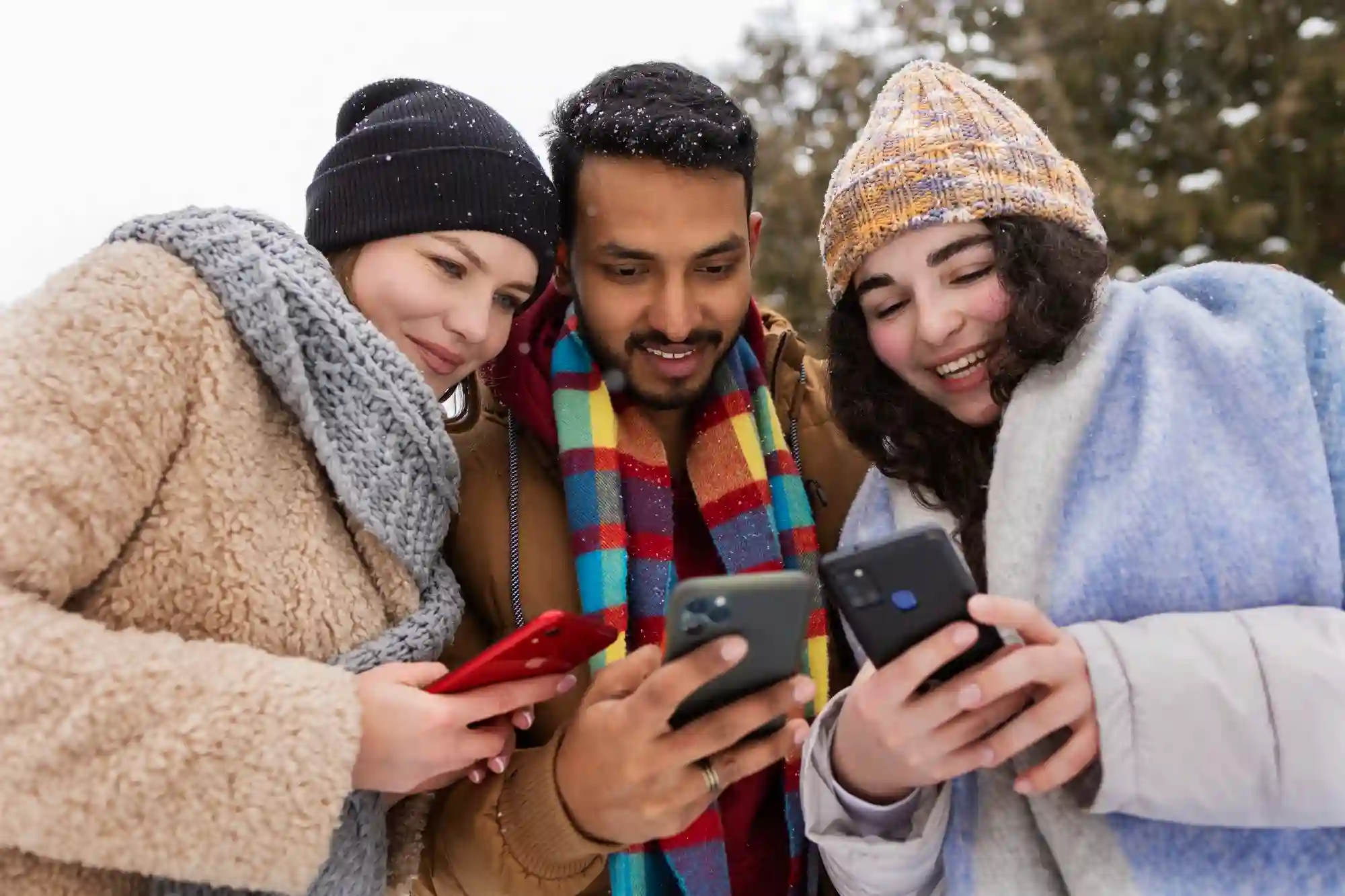 Three friends in winter gear smile while looking at their smartphones, sharing music and digital content.