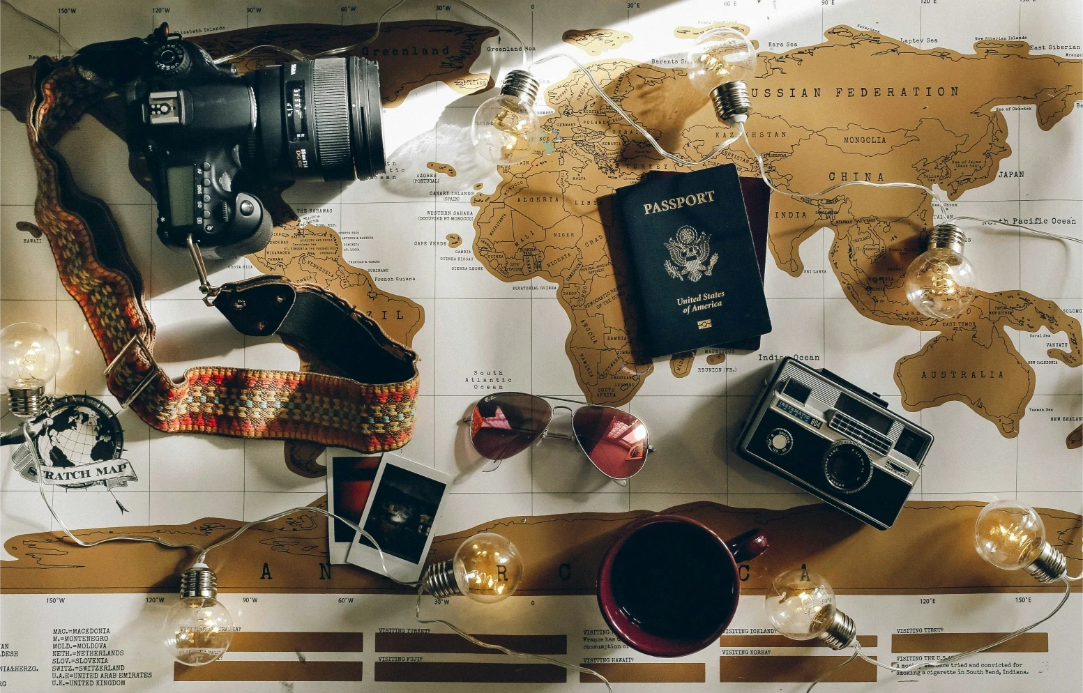 A flat lay of a world map with a camera, passport, sunglasses, retro Polaroid, and string lights, conveying a sense of travel and adventure.