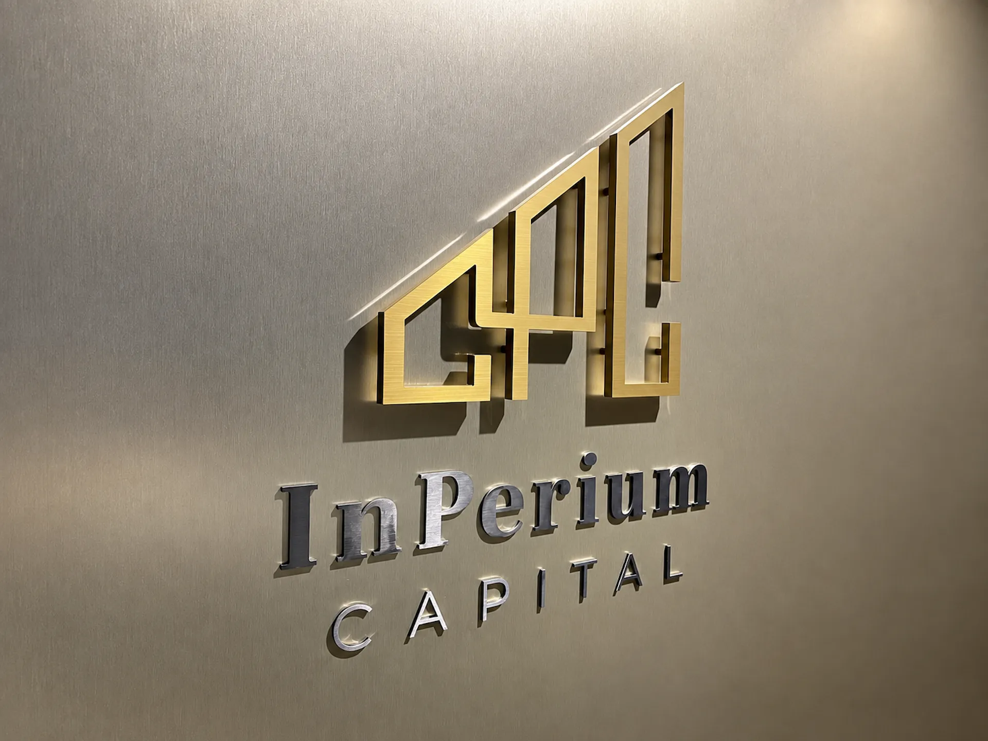 Zlaté 3D logo a stříbrný nápis InPerium Capital na světlé stěně v interiéru kanceláře
