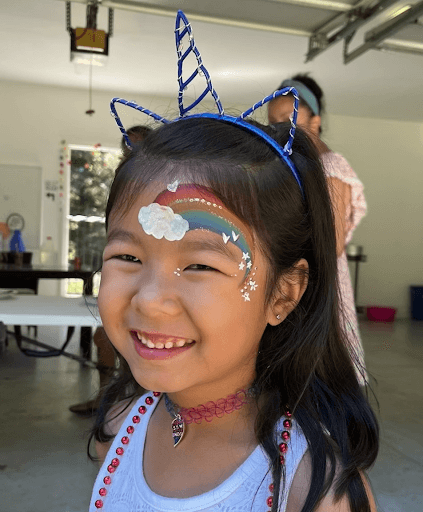 Smiling young girl with rainbow and cloud face paint, wearing sparkly unicorn ear and horn headbands, in a brightly lit indoor setting with people in the background