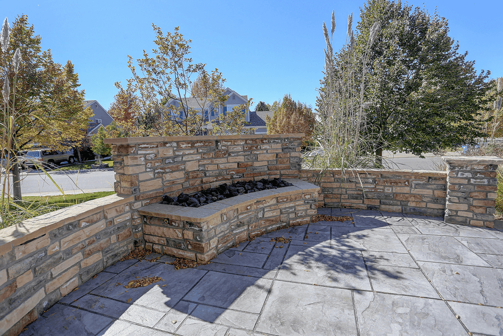 A curved stone outdoor fireplace bench set on a paved patio, surrounded by seasonal grasses and trees in a suburban neighborhood. The fire pit is filled with black stones, adding a contemporary touch to the rustic stonework.