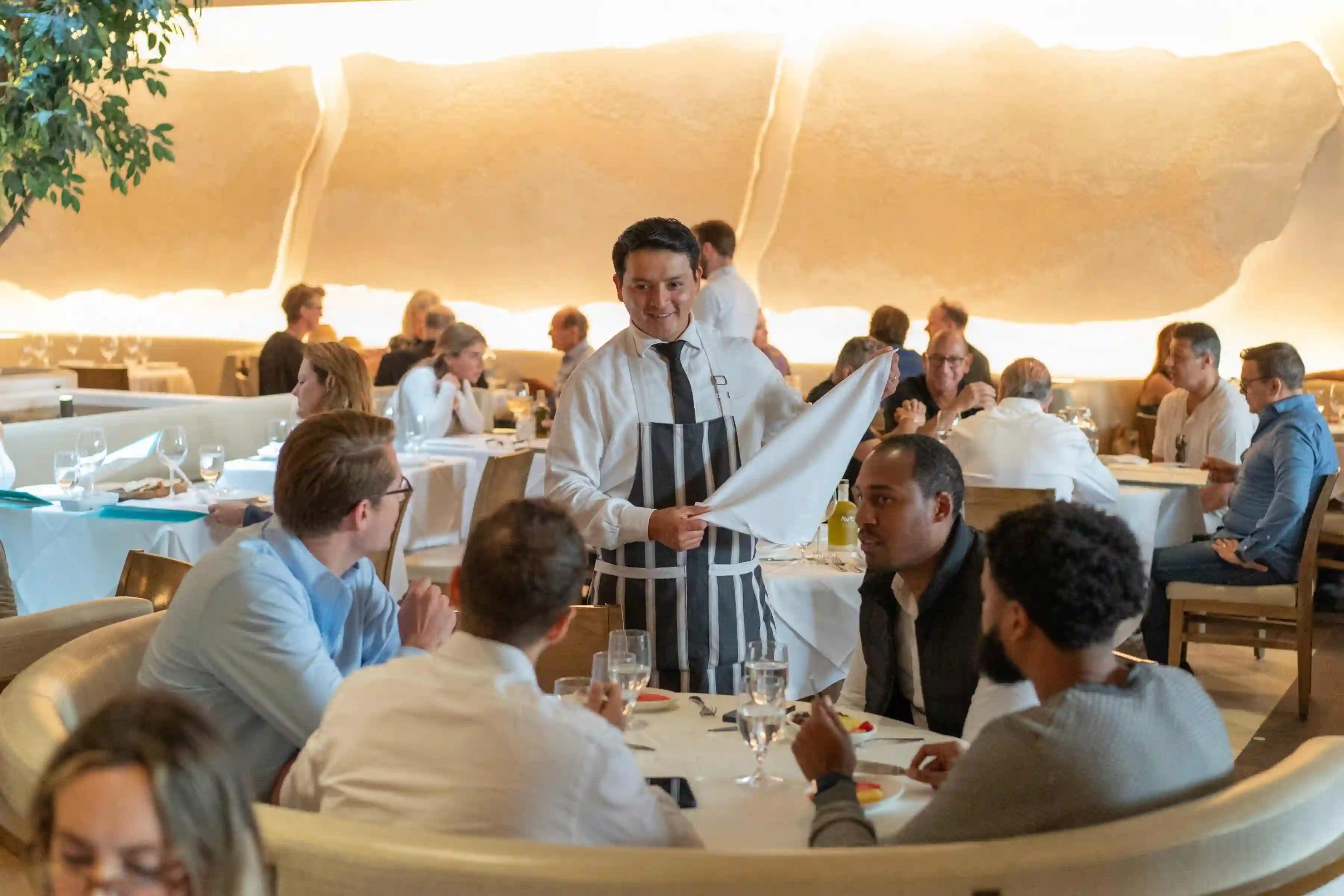 A waiter serves at a busy restaurant, where patrons are engaged in conversation at elegantly set tables.