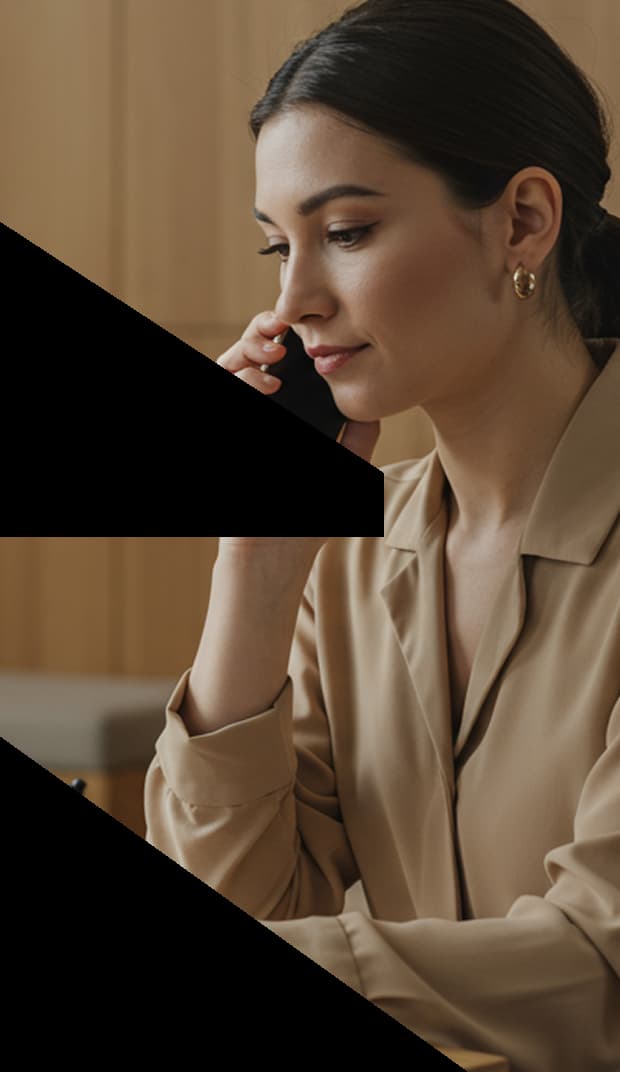 The image shows a woman sitting at a table, talking on a smartphone. She is wearing a beige blouse and gold hoop earrings, holding a pen in her other hand as if she’s taking notes. The background features light wooden panels, creating a calm and professional atmosphere.