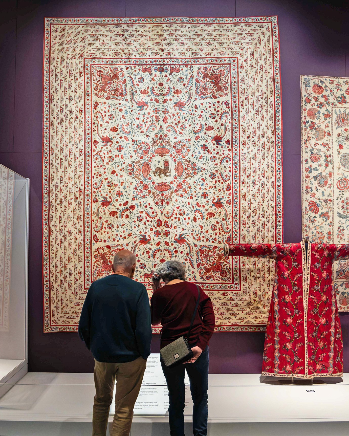 A couple stand in front of a very large gold and red textile hanging on an exhibition wall